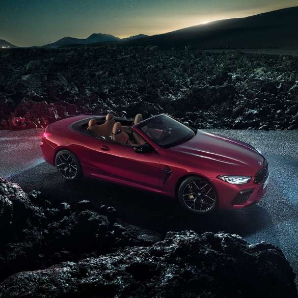 BMW M8 Competition Cabrio, tročetvrtinski bočni prikaz. BMW M8 Competition Cabrio u Motegi Red metalik boji s otvorenim mekim krovom, tročetvrtinski bočni prikaz, na asfaltu noću.
