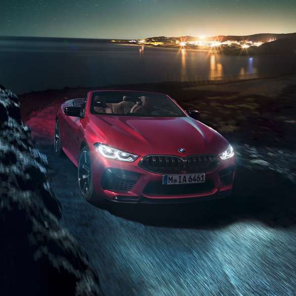 BMW M8 Competition Cabrio, tročetvrtinski prikaz prednjeg kraja. BMW M8 Competition Cabrio u Motegi Red metalik boji s otvorenim mekim krovom, tročetvrtinski prikaz prednjeg kraja, u vožnji noću na obalnoj cesti.