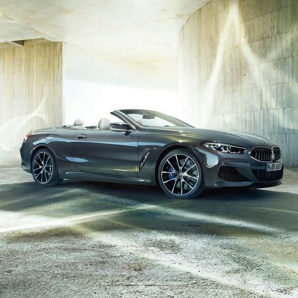 BMW M850i xDrive Cabrio, tročetvrtinski bočni prikaz. BMW M850i xDrive Cabrio u Dravit Grey metalik boji, sa spuštenim mekim krovom, tročetvrtinski bočni prikaz, ispred betonske zgrade.