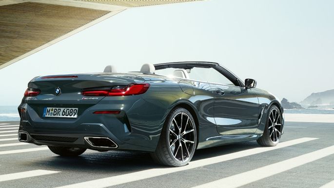 BMW M850i xDrive Cabrio, tročetvrtinski prikaz stražnjeg kraja. BMW M850i xDrive Cabrio u Dravit Grey metalik boji, sa spuštenim mekim krovom, tročetvrtinski prikaz stražnjeg kraja, parkiran s pogledom na ocean.