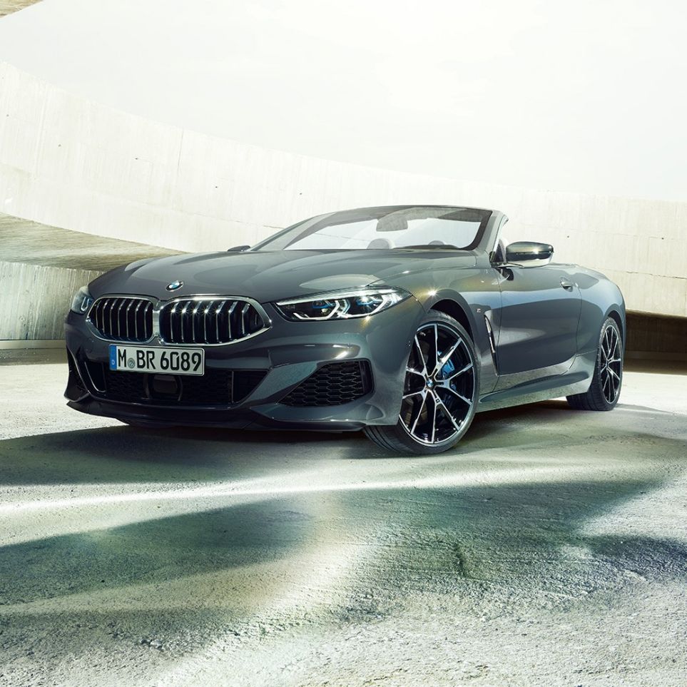 BMW M850i xDrive Cabrio, tročetvrtinski prikaz prednjeg kraja. BMW M850i xDrive Cabrio u Dravit Grey metalik boji, sa spuštenim mekim krovom, tročetvrtinski prikaz prednjeg kraja, ispred betonske zgrade.