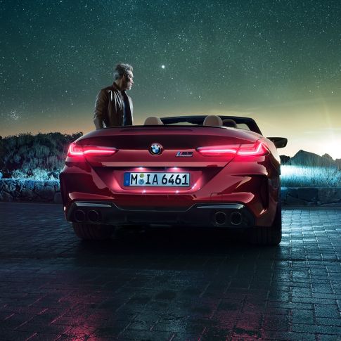BMW M8 Competition Cabrio, prikaz stražnjeg kraja. BMW M8 Competition Cabrio u Motegi Red metalik boji sa spuštenim mekim krovom, prikazom stražnjeg kraja, s čovjekom pod zvjezdanim nebom.