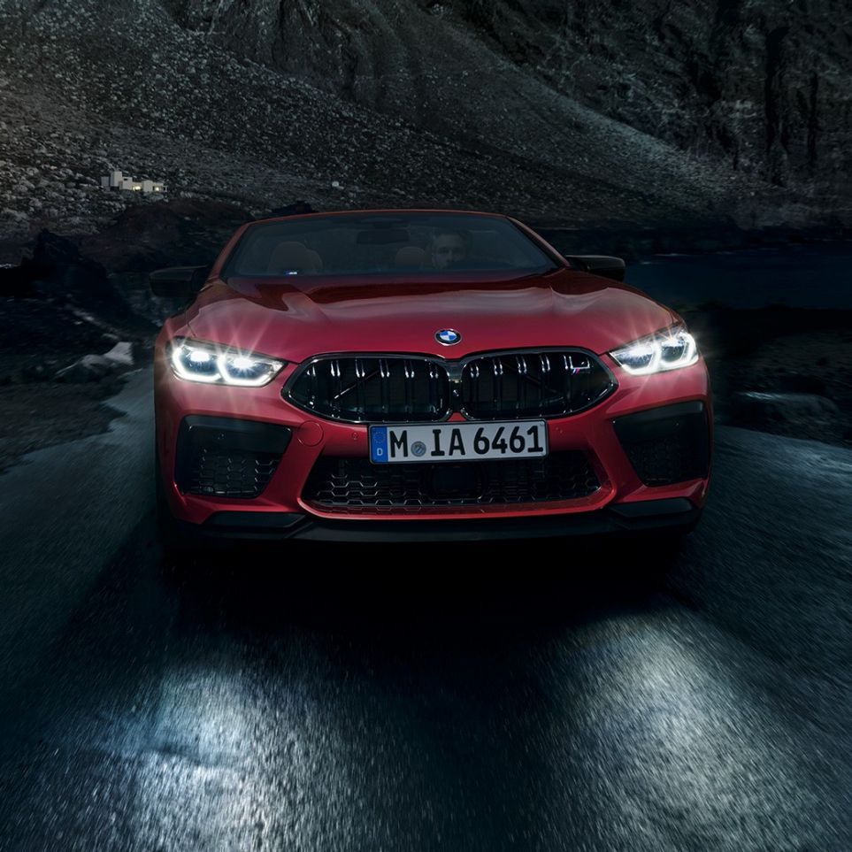 BMW M8 Competition Cabrio, prikaz prednjeg kraja. BMW M8 Competition Cabrio u Motegi Red metalik boji sa sjajnim prednjim svjetlima, prikaz prednjeg kraja, u vožnji noću.