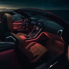 BMW M8 Competition Cabrio, unutrašnjost. BMW M8 Competition Cabrio u Motegi Red metalik boji sa spuštenim mekim krovom, unutrašnjost s crvenim ambijentalnim svjetlom noću.
