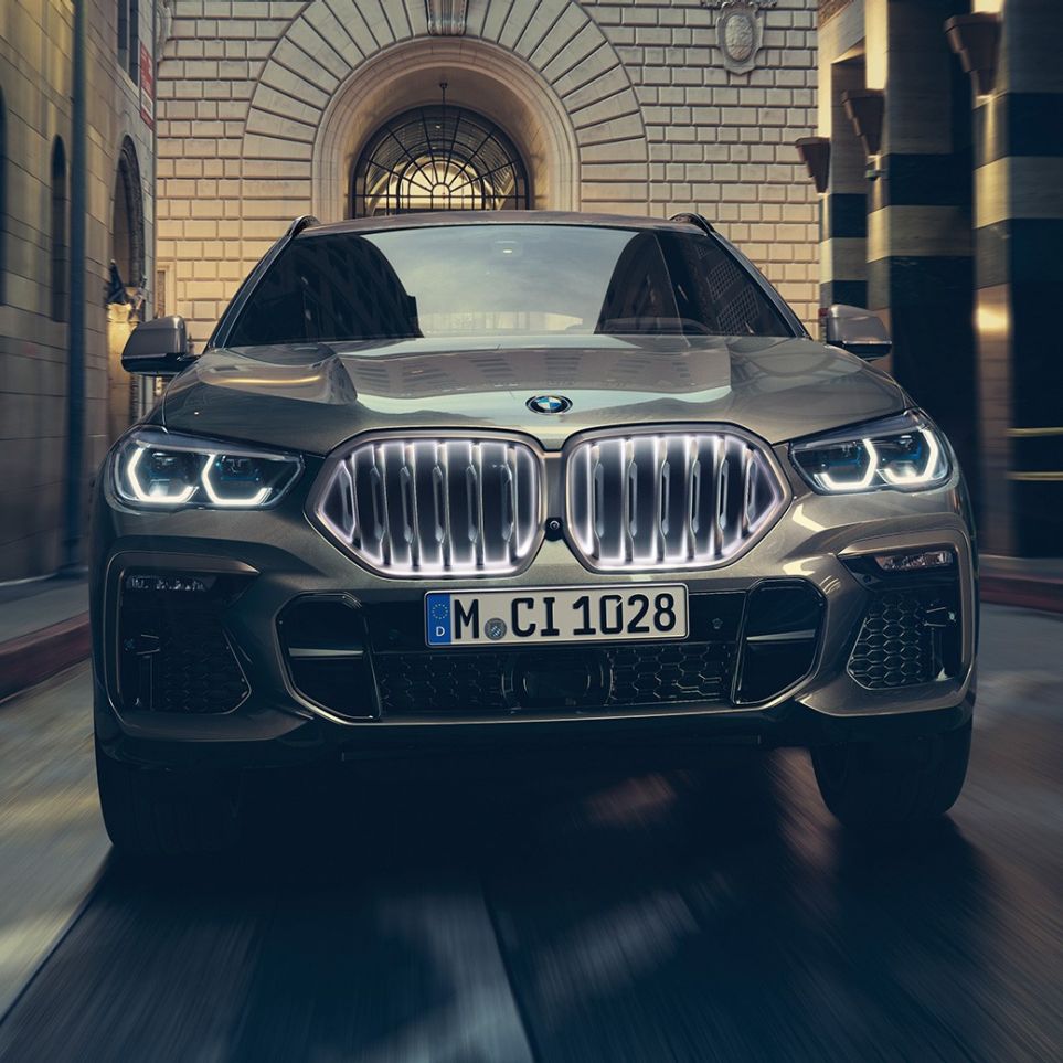 BMW X6 s BMW ‘Iconic Glow’ rešetkom maske. Prikaz prednjeg kraja BMW-a X6 ispred urbanog okruženja.