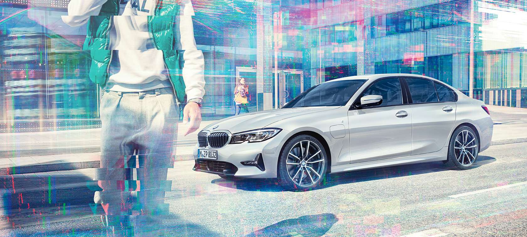 BMW serije 3 limuzina, bočni prikaz BMW-a 330e. Tročetvrtinski bočni prikaz BMW-a 330e limuzina ispred urbane staklene zgrade s putnicima koji hodaju.