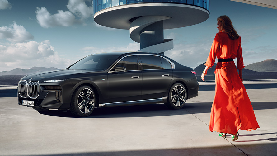 Model u crvenoj haljini ispred BMW serije 7 Limuzina G70 ICE M sport paket  BMW serije 7 Limuzina G70 ICE M sport paket Frozen Deep grey metalik 21" M naplatci od lakih legura star spoke 908 tročetvrtinski prednji pogled s modelom u crvenoj haljini