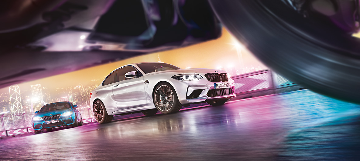 BMW M2 Competition na asfaltu. Dva modela BMW M2 Competition na trkaćoj stazi ispred noćnog pejzaža grada.