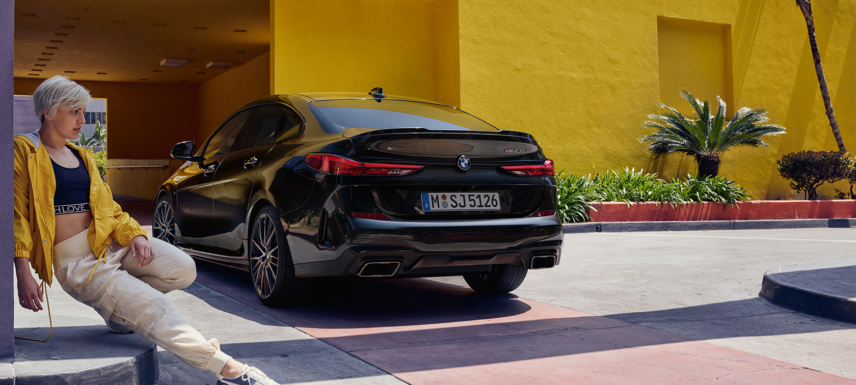 BMW M235i xDrive Gran Coupé tročetvrtinski stražnji pogled BMW M235i xDrive Gran Coupé Black Sapphire metalik F44 tročetvrtinski stražnji pogled sa ženom ispred automobila