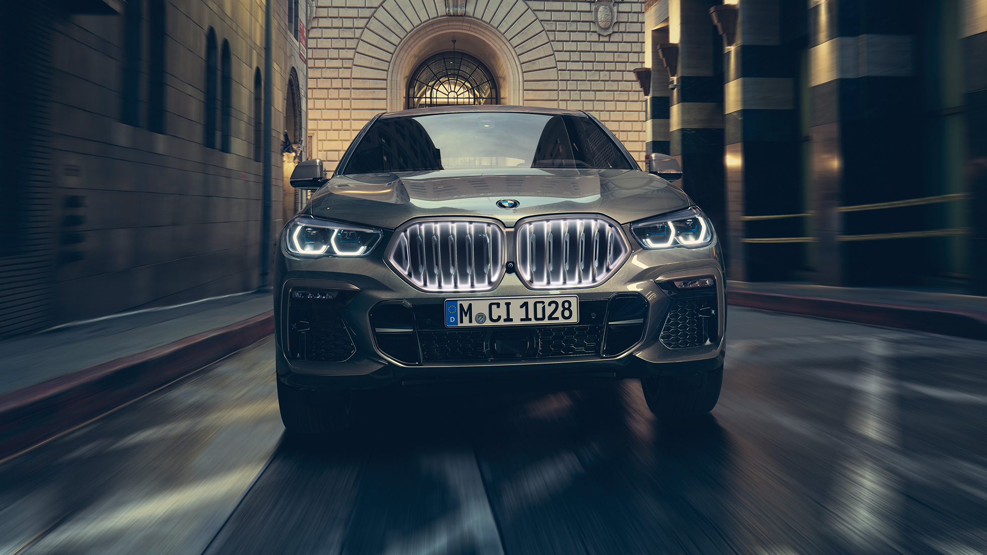 BMW X6 M50i s BMW ‘Iconic Glow‘ rešetkom maske. Prikaz prednjeg kraja BMW-a X6 M50i ispred urbanog okruženja.