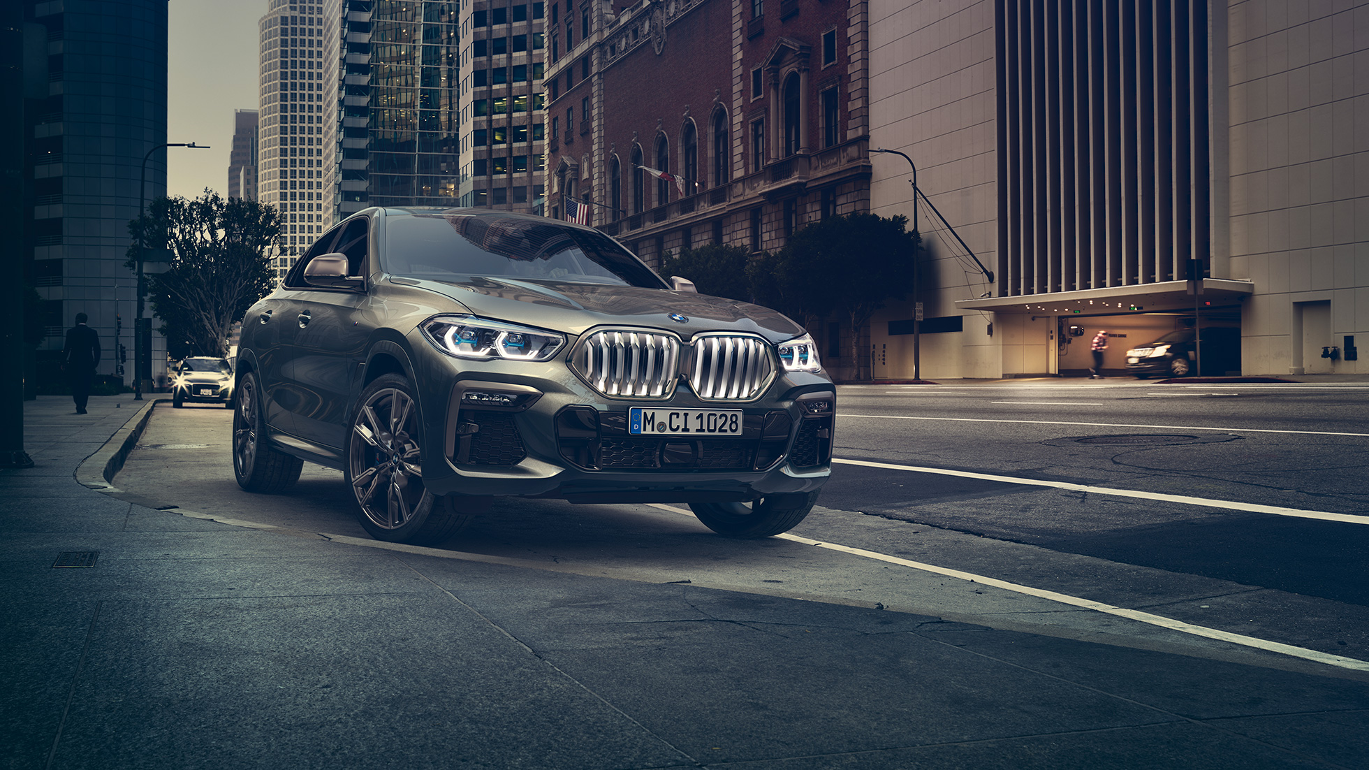 BMW X6 M50i u stacionarnom položaju. BMW X6 M50i u tro-četvrtinskom prikazu prednjeg kraja stoji ispred urbanog okruženja.