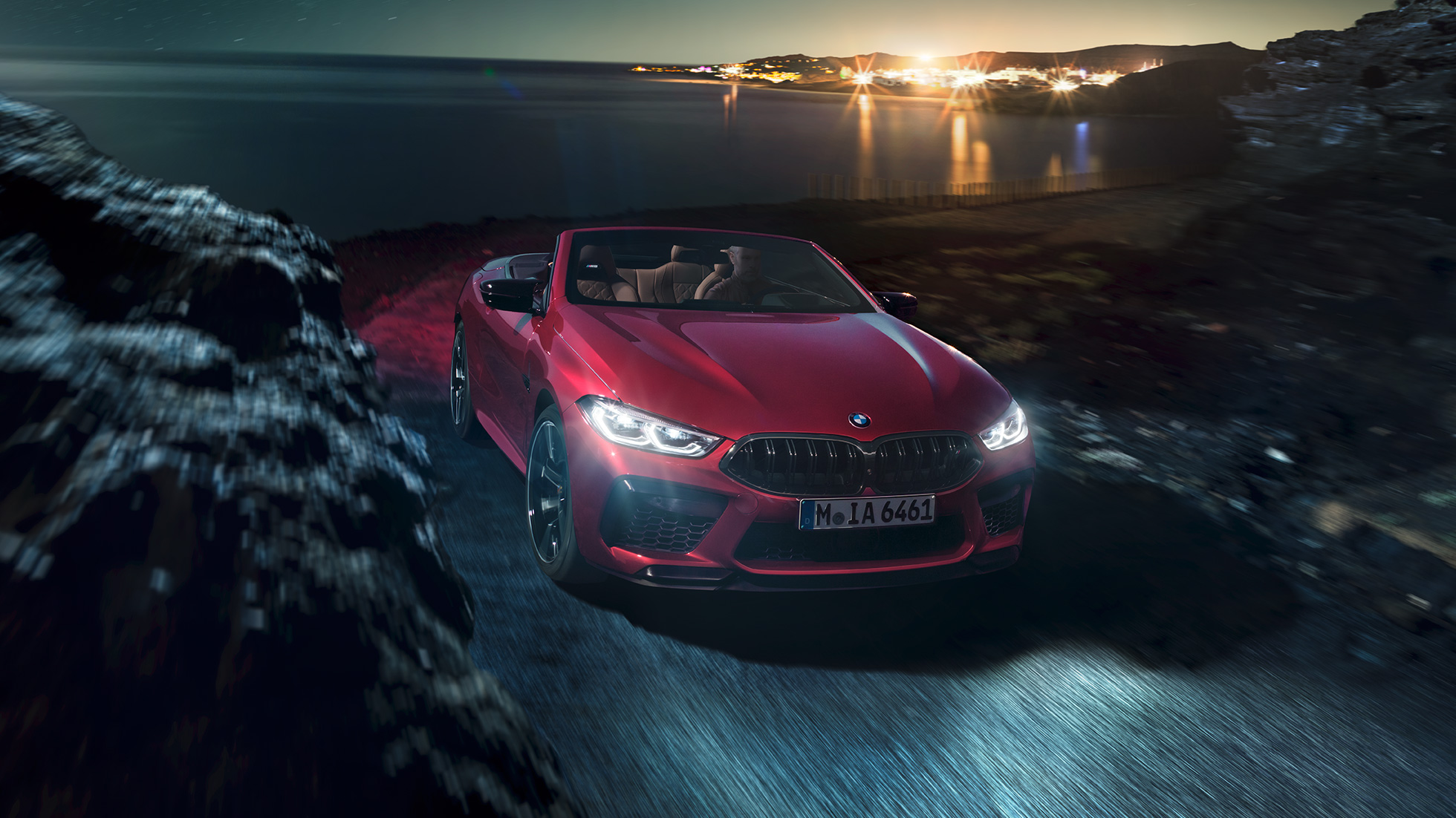 BMW M8 Competition Cabrio, tročetvrtinski prikaz prednjeg kraja. BMW M8 Competition Cabrio u Motegi Red metalik boji s otvorenim mekim krovom, tročetvrtinski prikaz prednjeg kraja, u vožnji noću na obalnoj cesti.