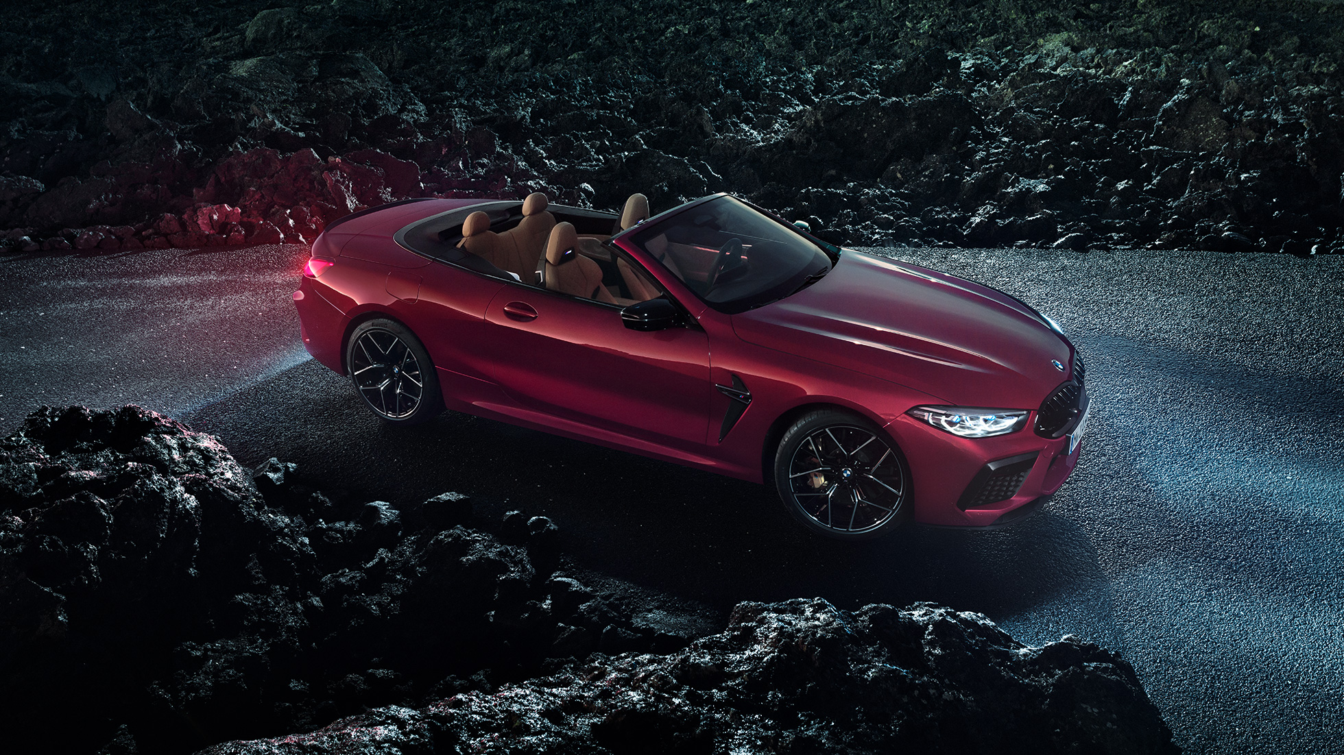 BMW M8 Competition Cabrio, tročetvrtinski bočni prikaz. BMW M8 Competition Cabrio u Motegi Red metalik boji s otvorenim mekim krovom, tročetvrtinski bočni prikaz, na asfaltu noću.