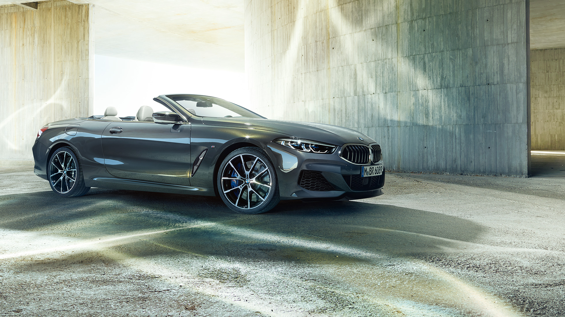 BMW M850i xDrive Cabrio, tročetvrtinski bočni prikaz. BMW M850i xDrive Cabrio u Dravit Grey metalik boji, sa spuštenim mekim krovom, tročetvrtinski bočni prikaz, ispred betonske zgrade.
