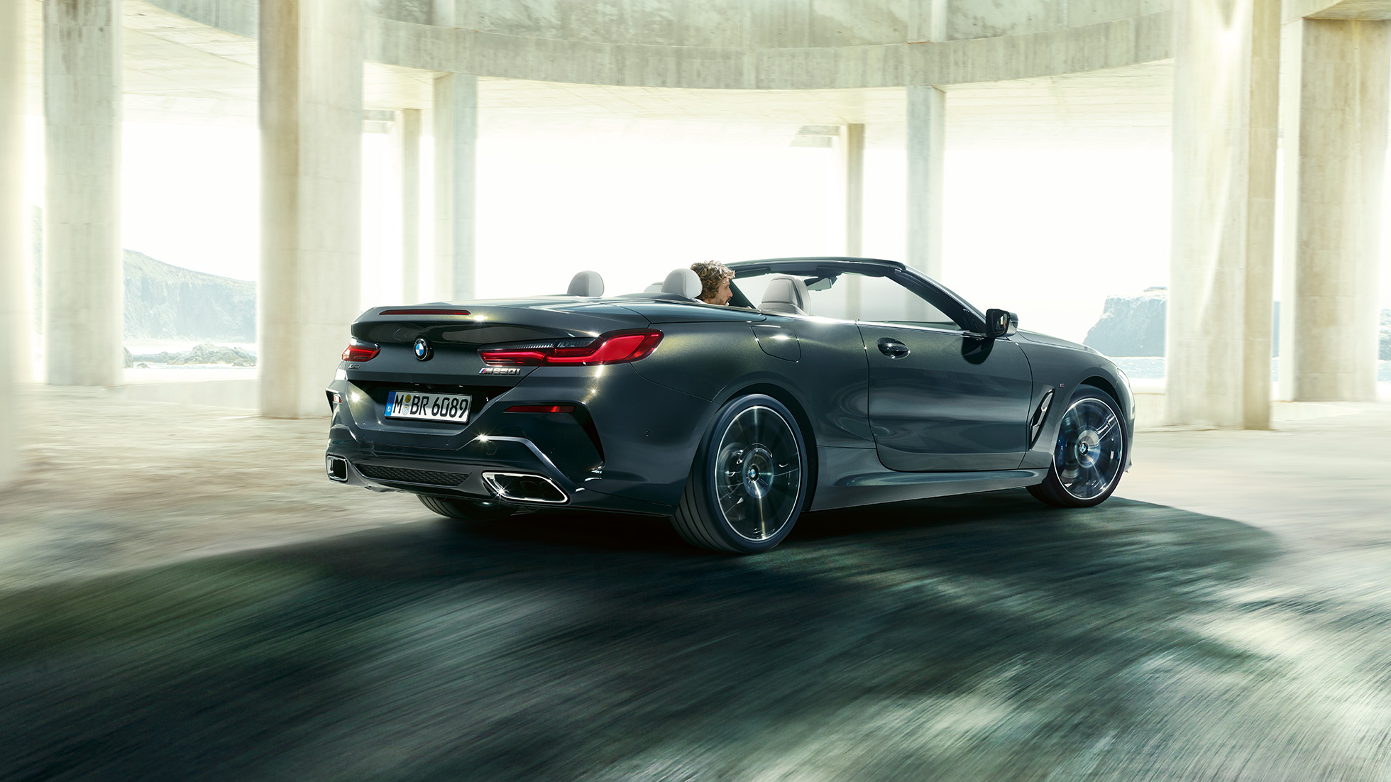 BMW M850i xDrive Cabrio, tročetvrtinski prikaz stražnjeg kraja. BMW M850i xDrive Cabrio u Dravit Grey metalik boji, sa spuštenim mekim krovom, tročetvrtinski prikaz stražnjeg kraja, u vožnji kroz betonsku zgradu.