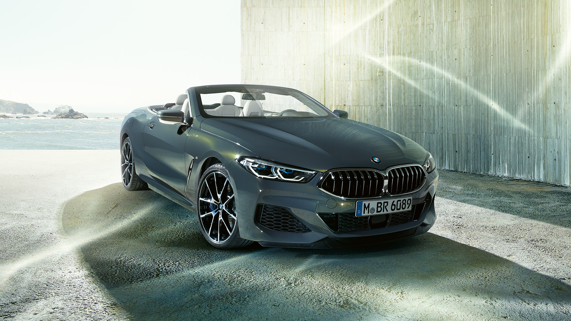 BMW M850i xDrive Cabrio, tročetvrtinski prikaz prednjeg kraja. BMW M850i xDrive Cabrio u Dravit Grey metalik boji, sa spuštenim mekim krovom, tročetvrtinski prikaz prednjeg kraja, ispred obale.