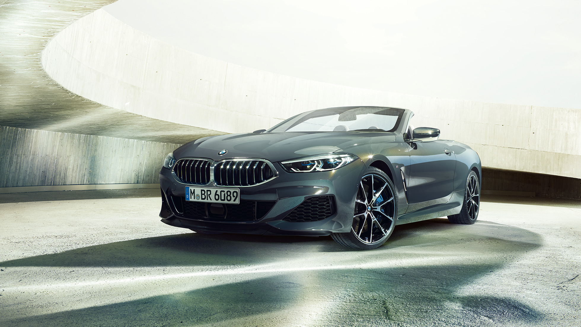 BMW M850i xDrive Cabrio, tročetvrtinski prikaz prednjeg kraja. BMW M850i xDrive Cabrio u Dravit Grey metalik boji, sa spuštenim mekim krovom, tročetvrtinski prikaz prednjeg kraja, ispred betonske zgrade.