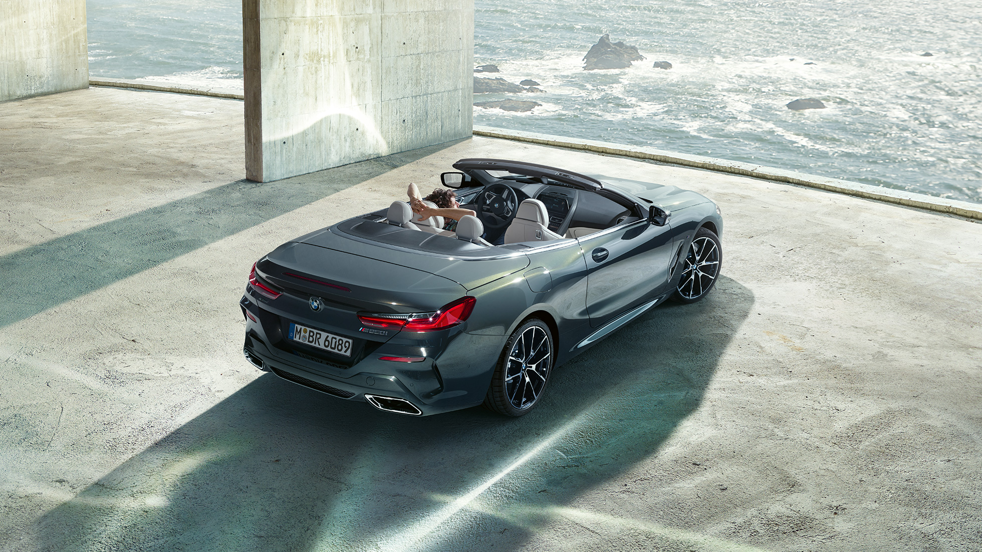 BMW M850i xDrive Cabrio, prikaz iz ptičje perspektive. BMW M850i xDrive Cabrio u Dravit Grey metalik boji, sa spuštenim mekim krovom, prikaz iz ptičje perspektive, parkiran u betonskoj zgradi ispred oceana.