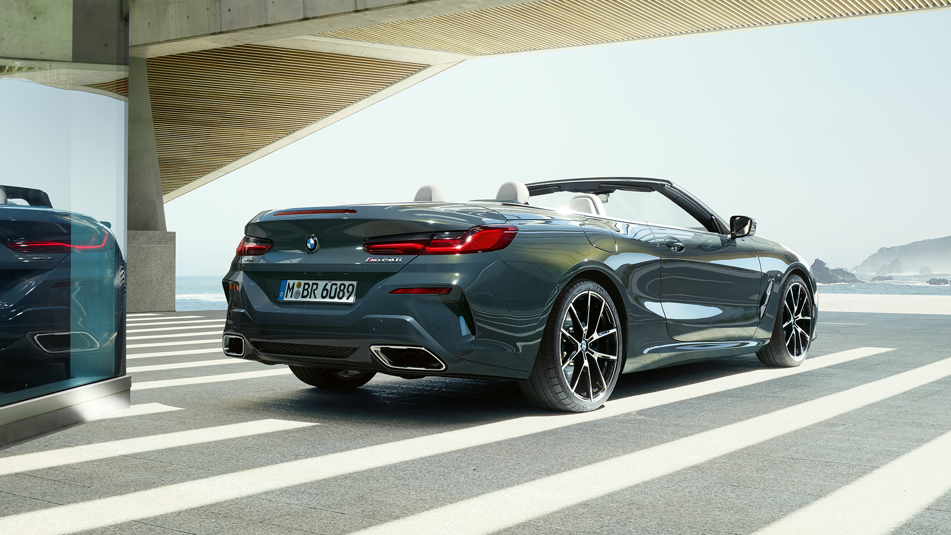 BMW M850i xDrive Cabrio, tročetvrtinski prikaz stražnjeg kraja. BMW M850i xDrive Cabrio u Dravit Grey metalik boji, sa spuštenim mekim krovom, tročetvrtinski prikaz stražnjeg kraja, parkiran s pogledom na ocean.