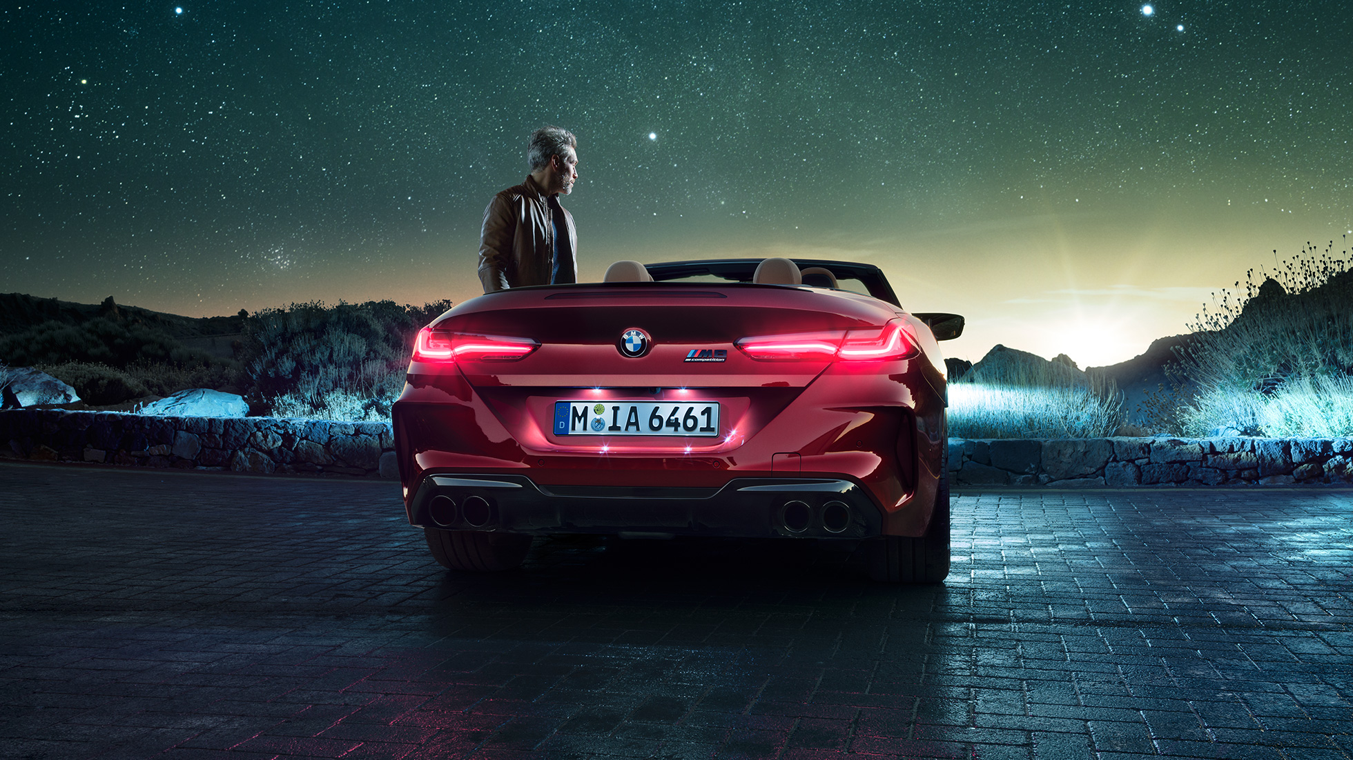 BMW M8 Competition Cabrio, prikaz stražnjeg kraja. BMW M8 Competition Cabrio u Motegi Red metalik boji sa spuštenim mekim krovom, prikazom stražnjeg kraja, s čovjekom pod zvjezdanim nebom.