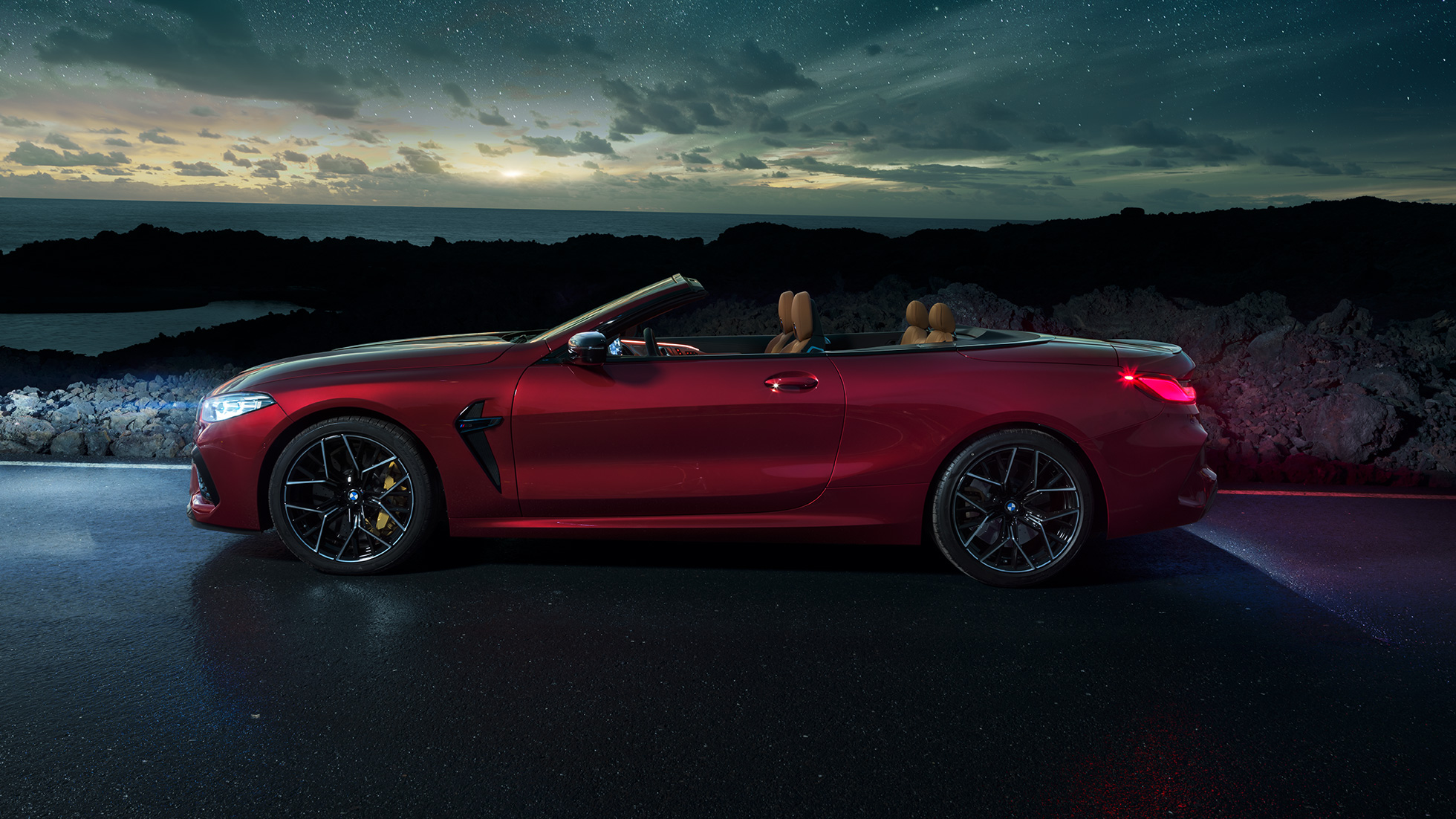 BMW M8 Competition Cabrio, bočni prikaz. BMW M8 Competition Cabrio u Motegi Red metalik boji sa spuštenim mekim krovom, bočni prikaz, noću pod zvjezdanim nebom.