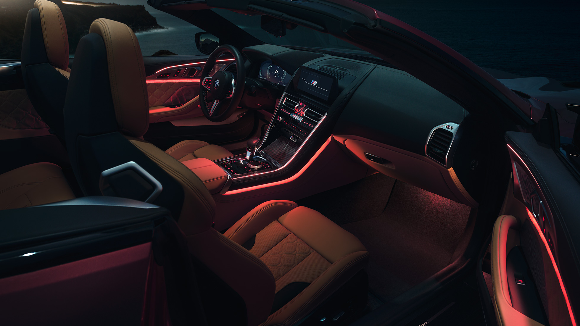 BMW M8 Competition Cabrio, unutrašnjost. BMW M8 Competition Cabrio u Motegi Red metalik boji sa spuštenim mekim krovom, unutrašnjost s crvenim ambijentalnim svjetlom noću.