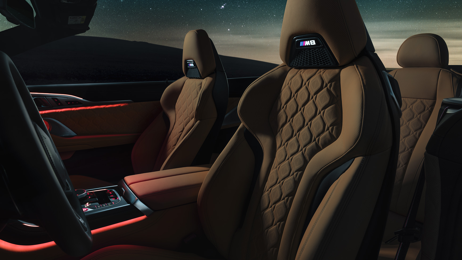 M Sport sjedala u 'Merino' koži. BMW M8 Competition Cabrio sa spuštenim mekim krovom pod zvjezdanim nebom, unutrašnjost s M Sport sjedalima u 'Merino' koži i crvenim ambijentalnim svjetlom.