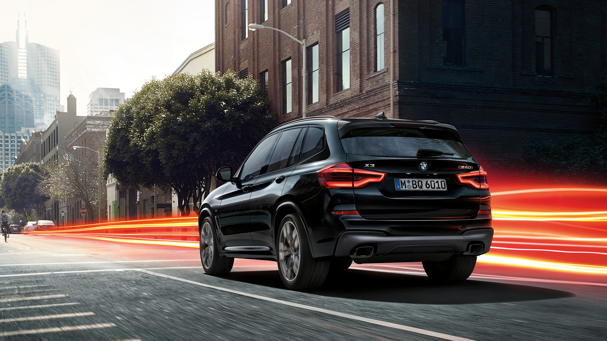 BMW X3 M40i, tročetvrtinski stražnji prikaz. BMW X3 M40i u Black Sapphire metalik boji, u vožnji, vanjština, tročetvrtinski stražnji prikaz ispred urbane pozadine sa svijetlosnim trakama.