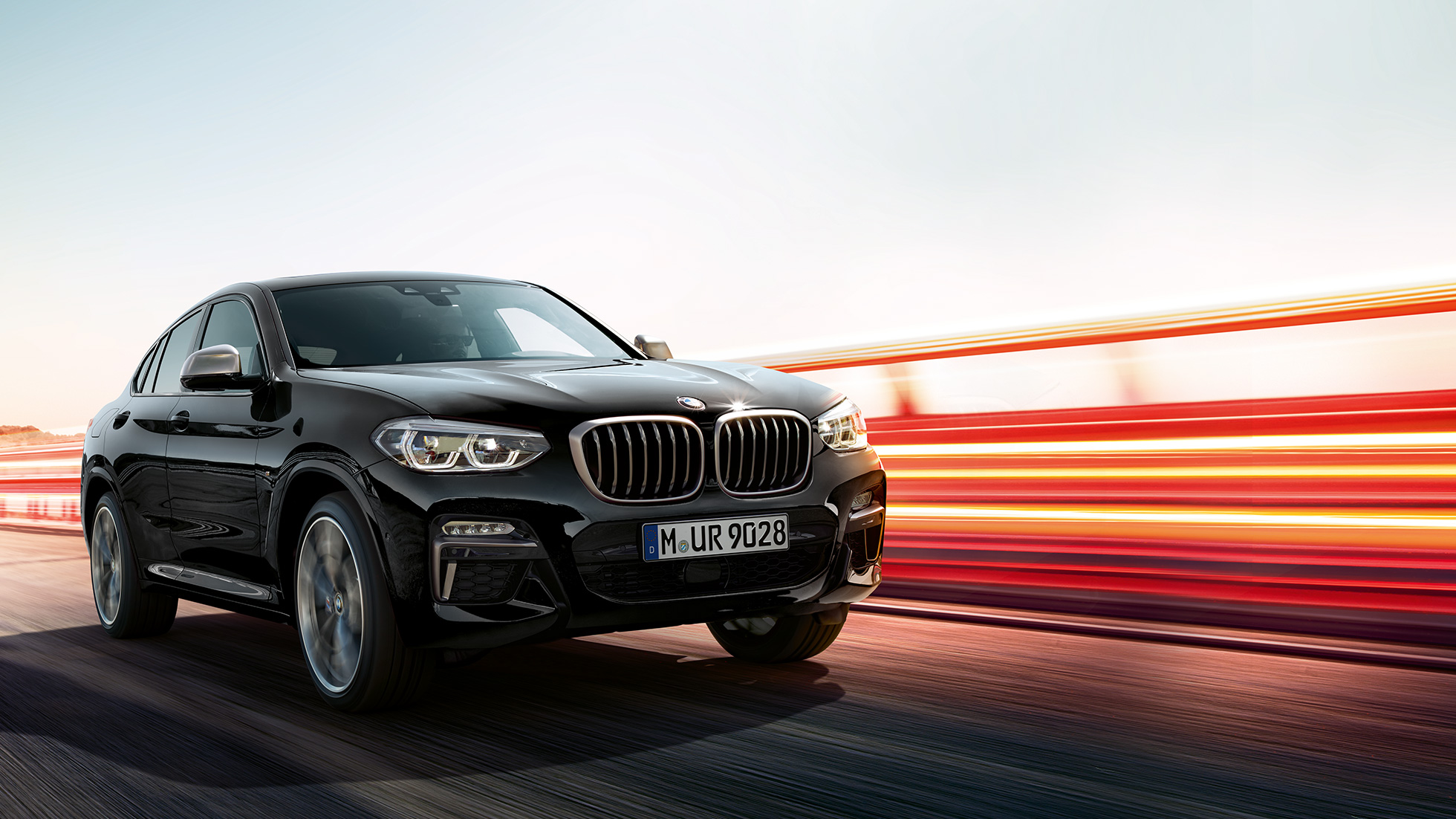BMW X4 M40i, tročetvrtinski stražnji prikaz. BMW X4 M40i u Black Sapphire metalik boji, u vožnji, vanjština, tročetvrtinski stražnji prikaz sa svijetlosnim trakama.