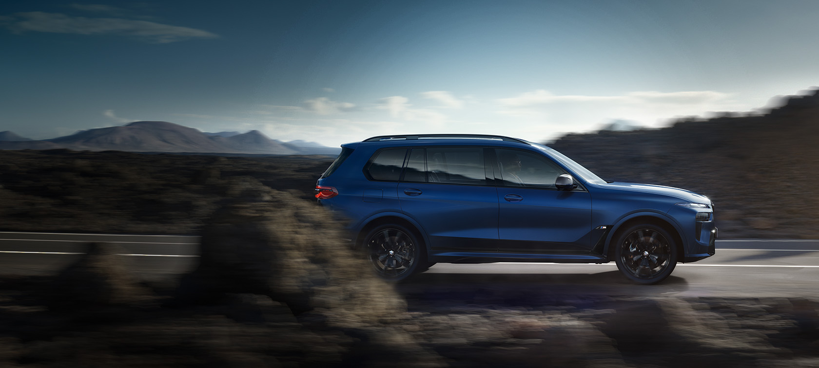 BMW X7 M60i xDrive G07 LCI Frozen Marina Bay Blue bočni pogled u vožnji na ulici ispred planinskog krajolika BMW X7 M60i xDrive G07 LCI Frozen Marina Bay Blue bočni pogled u vožnji na ulici