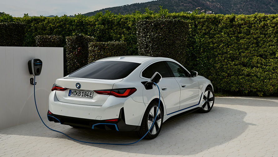 Tročetvrtinski prikaz stražnjeg kraja BMW-a i4 G26 BEV prilikom punjenja spojen na BMW Wallbox Gen 3 BMW i4 G26 BEV tročetvrtinski prikaz stražnjeg kraja prilikom punjenja spojen na BMW Wallbox Gen 3 ispred živice