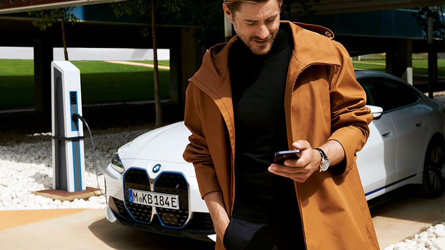 Trajanje paketa BMW Service Inclusive i ugovorna razdoblja za električna vozila BMW Service Inclusive muškarac gleda u pametni telefon ispred BMW-a i4 na stanici za punjenje